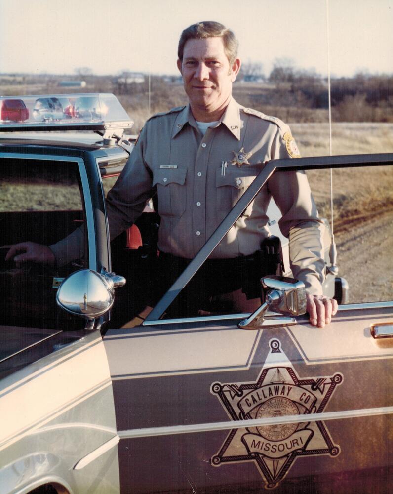 A color image of Harry Lee standing by the driver's side of a patrol vehicle.